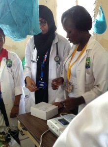 Ruth Oenga (right) and Sakina Abdallah (partially obscured by bednet) using EKG machine with a hospital patient.