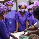 Midwives dressed in purple scrubs leaning at the bedside in a clinical setting.